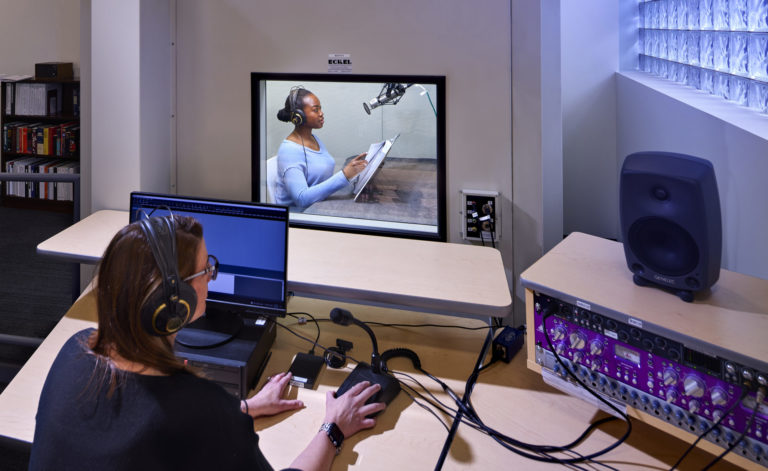 Woman in the foreground runs a soundboard to record a volunteer who reads a book aloud.