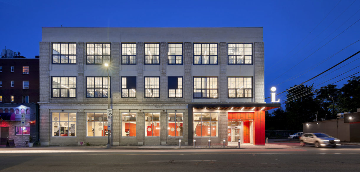 Light and activity shine out of the windows of a newly-renovated building. Bold red tile marks a recessed, corner entrance.